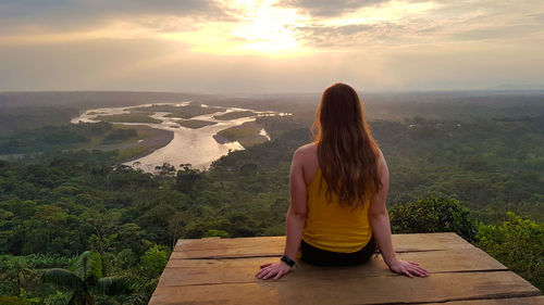 Rear view of woman sitting on landscape against sunset sky