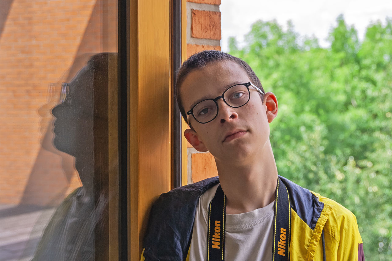 headshot, one person, portrait, eyeglasses, young adult, front view, glasses, young men, real people, window, architecture, day, lifestyles, building exterior, looking away, looking, standing, leisure activity, outdoors, teenage boys, contemplation, teenager