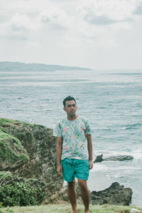Full length portrait of young man standing on rock against sea