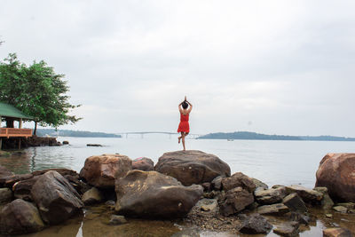 Rear view of woman doing yoga on rock by sea against sky