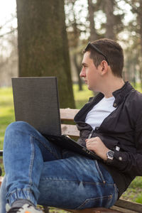 Young man sitting with laptop on bench at park