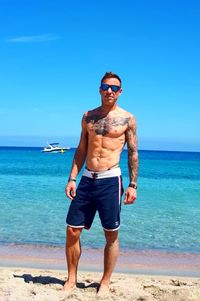 Full length of shirtless man standing at beach against blue sky