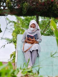 Full length of woman holding mobile phone while sitting outdoors