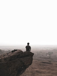 Rear view of man sitting on rock against clear sky