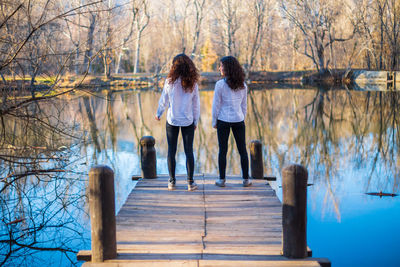 Full length of friends tossing hair while standing on pier by lake in forest