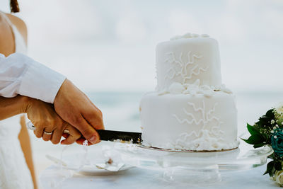 Cropped hands of newlywed couple cutting wedding cake against sea