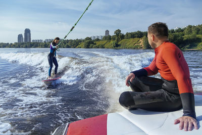 Man sitting at edge of moving boat looking at female wakeboarder surfing behind