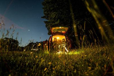 Unrecognizable friends sitting in illuminated camper van and enjoying freedom during camping in forest in summer night