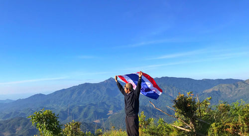 Man holding flag while standing by plants against mountains and blue sky