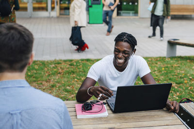 Young male student laughing while sitting with friend in campus