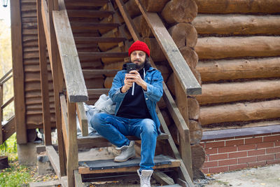 Man with a hipster beard in a red knitted hat and a denim jacket, is sit on stairs at wooden house 