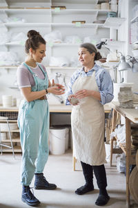 Senior female potter talking while working with young employee against shelves at workshop