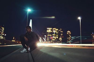 Full length of man sitting on roadside by light trails at night