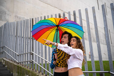 Young couple pointing at a point in amazement while holding an umbrella with pride colors.