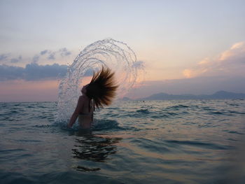 Side view of girl standing in sea with tousled hair splashing water in air against sky