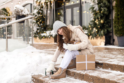 A happy girl in mittens sits on the steps and picks up gifts on the street in winter