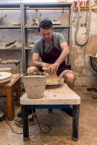 Full body of concentrated male master in apron sitting at table while sculpting with brown clay on throwing wheel
