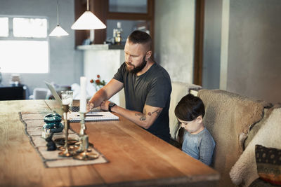 Man writing in book while son sitting at dining table