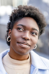 Thoughtful african american female with short hair looking up with smile while standing on street with buildings on blurred background