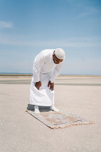 Full boy islamic male in traditional white clothes standing on rug and praying against blue sky on beach