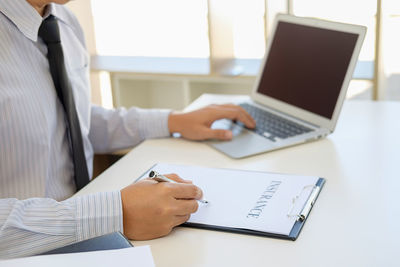 Midsection of insurance agent writing on paper while using laptop on table at office