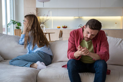 Frustrated spouses ignoring each other after quarrel manipulating silence sitting on sofa at home.