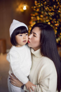 Beautiful korean mom and baby daughter sitting in white knitted sweaters at the christmas tree