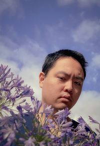 Low angle view of young man holding purple agapanthus lilies against cloudy blue sky.