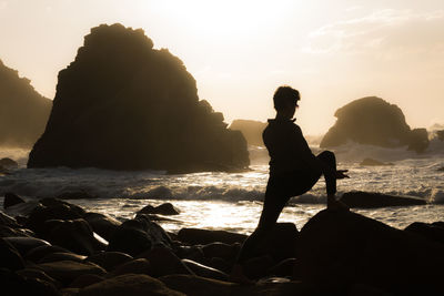 Silhouette woman doing yoga on rocks at beach during sunset