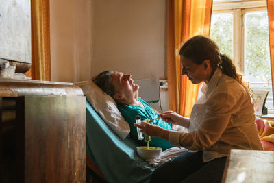 Side view of female nurse feeding paralyzed elderly lady with puree through tube and syringe while taking care of patient at home