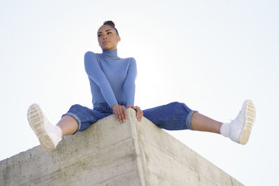 Thoughtful woman looking away while sitting on retaining wall against clear sky