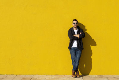 Young bearded man standing against a yellow wall with crossed arms