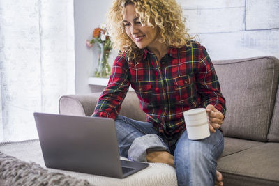 Smiling woman holding coffee cup using laptop at home