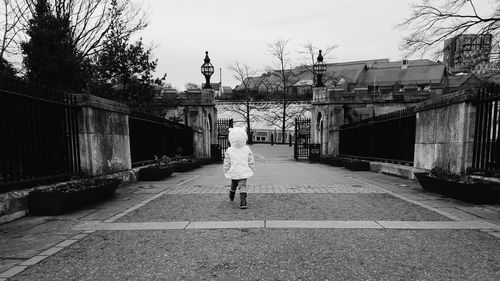 Rear view of girl walking on street by fence against sky