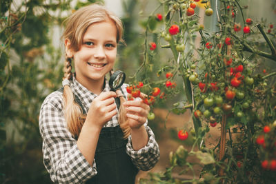 A beautiful little girl stands in a greenhouse with a crop, holds a magnifying glass in her hands
