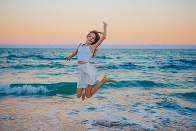 Full length of young woman with arms outstretched standing at beach against sky during sunset