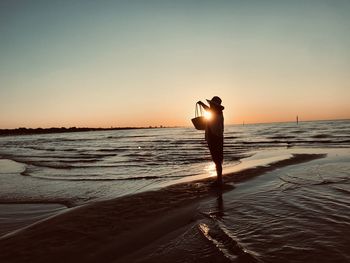 Silhouette woman holding bag standing on beach during sunset