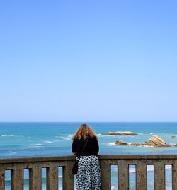 Rear view of woman leaning on railing against sea and clear sky