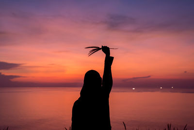 Rear view of silhouette woman holding plants while standing by sea against sky during sunset