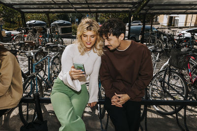Young woman sharing smart phone with male friend sitting on railing at college campus