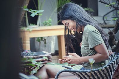 Side view of smiling young woman using mobile phone while sitting on chair in yard