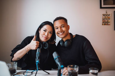 Portrait of smiling female blogger showing thumbs up with male colleague sitting against wall at home