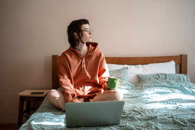 Thoughtful schoolgirl sitting on bed, looking at window during tea break after online studying