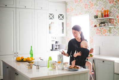 Mother using mobile phone while carrying daughter in kitchen at home