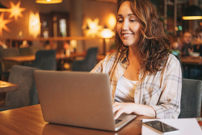 Adult charming brunette woman in plaid shirt working with laptop at cafe