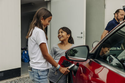 Smiling girl looking at sister charging electric car near house