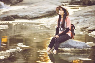 Thoughtful young woman wearing hat looking away while sitting on rock in lake