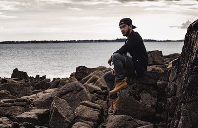 Young man sitting on cliff by sea against sky