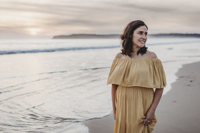 Lifestyle portrait of young woman standing on beach against cloudy sky