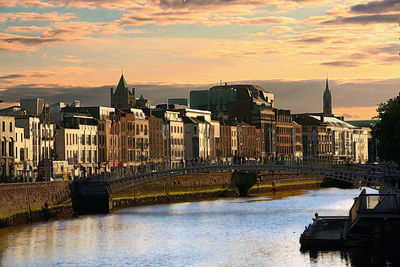 Bridge over river by buildings against sky during sunset